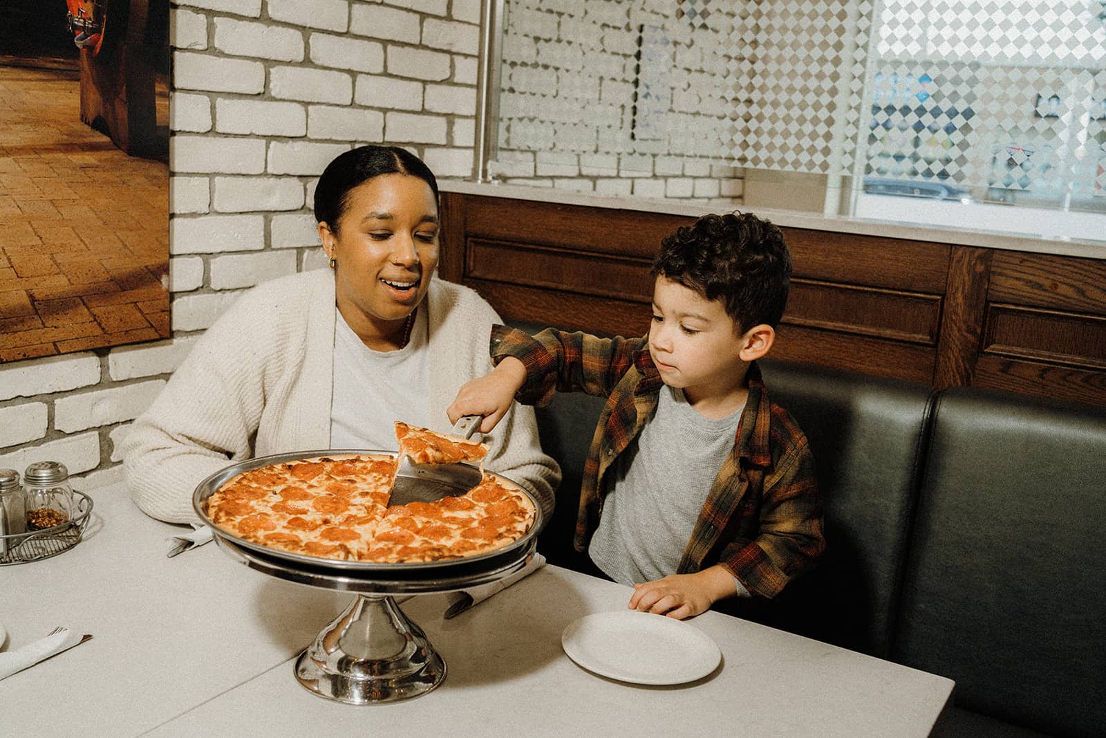 An adult and a child sit in a restaurant booth as the child serves a slice of pizza onto a plate.