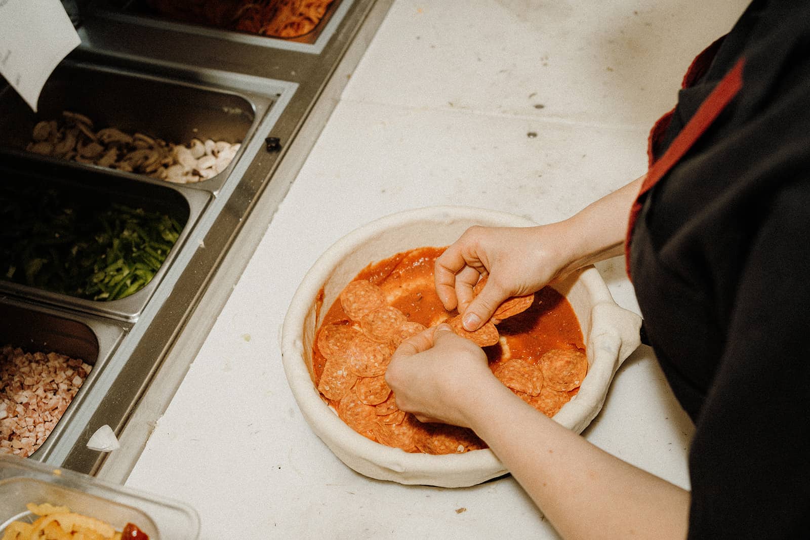 A person places pepperoni slices on a pizza crust with tomato sauce at a prep station with various toppings.