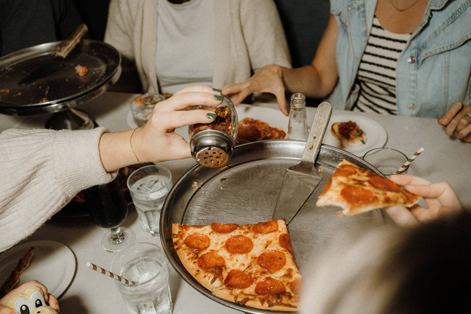 Four people sit at a table sharing pepperoni pizza; one hand sprinkles seasoning on a slice.