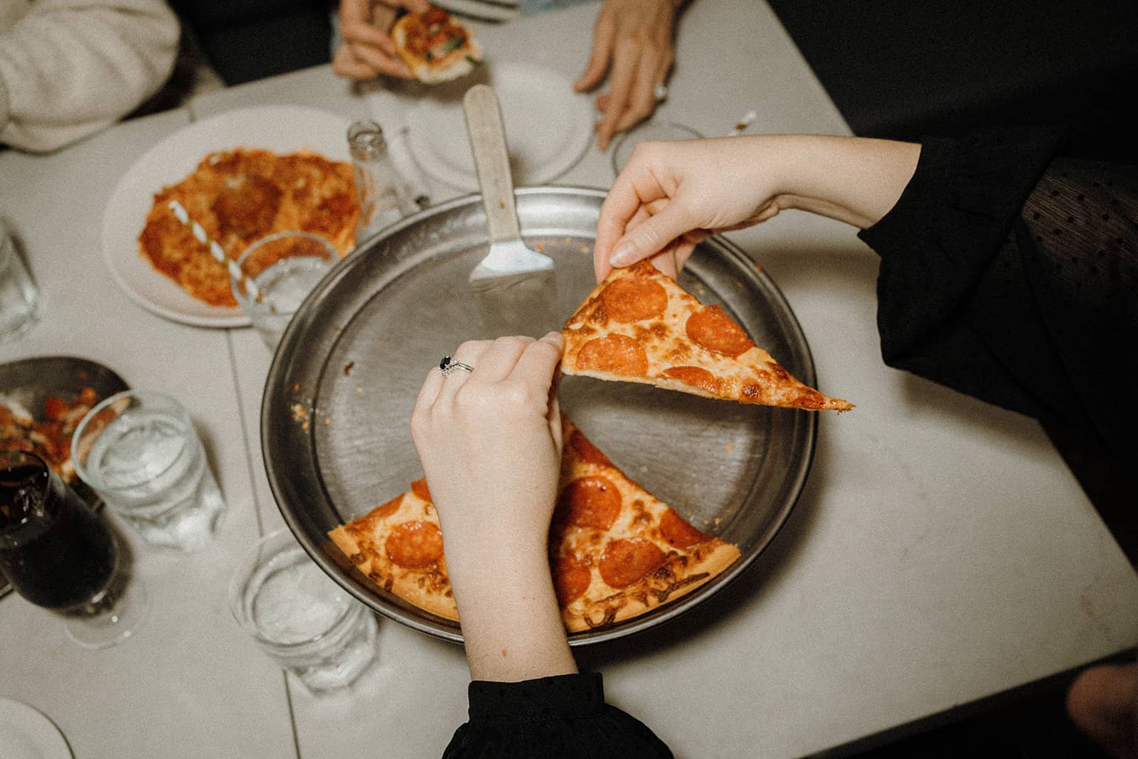 Two hands serve pepperoni pizza from a metal tray at a table set with drinks and more pizza.