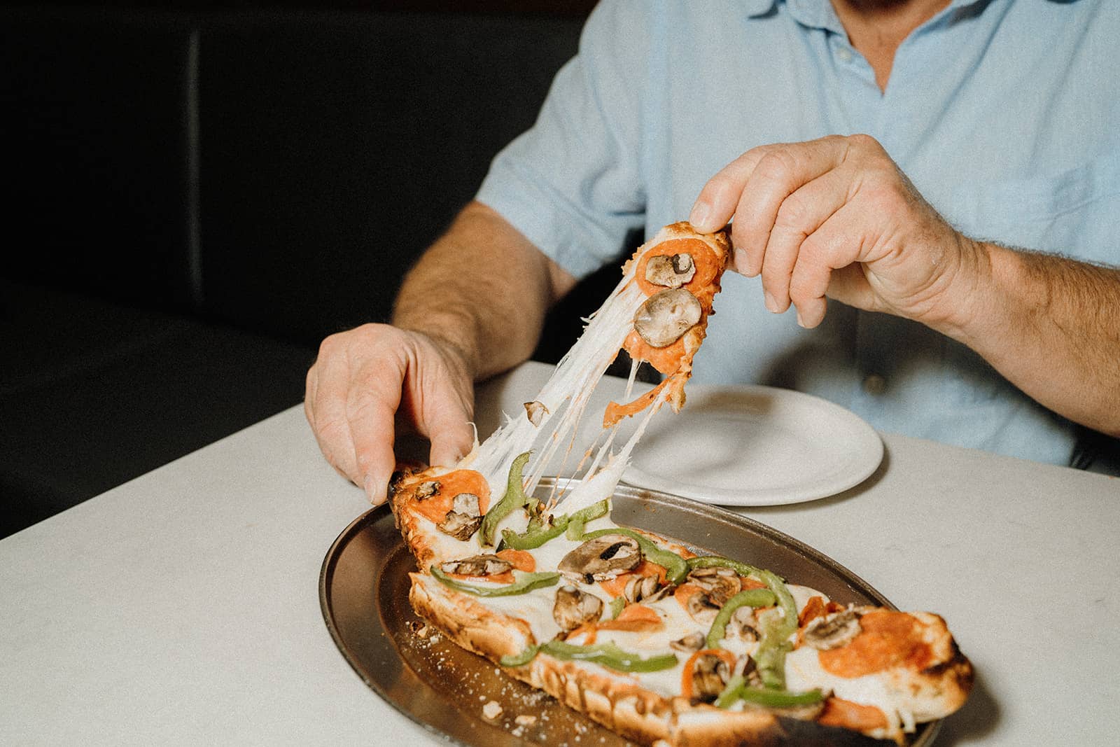 A person pulls a cheesy slice from a rectangular pizza topped with mushrooms and green peppers on a metal tray.